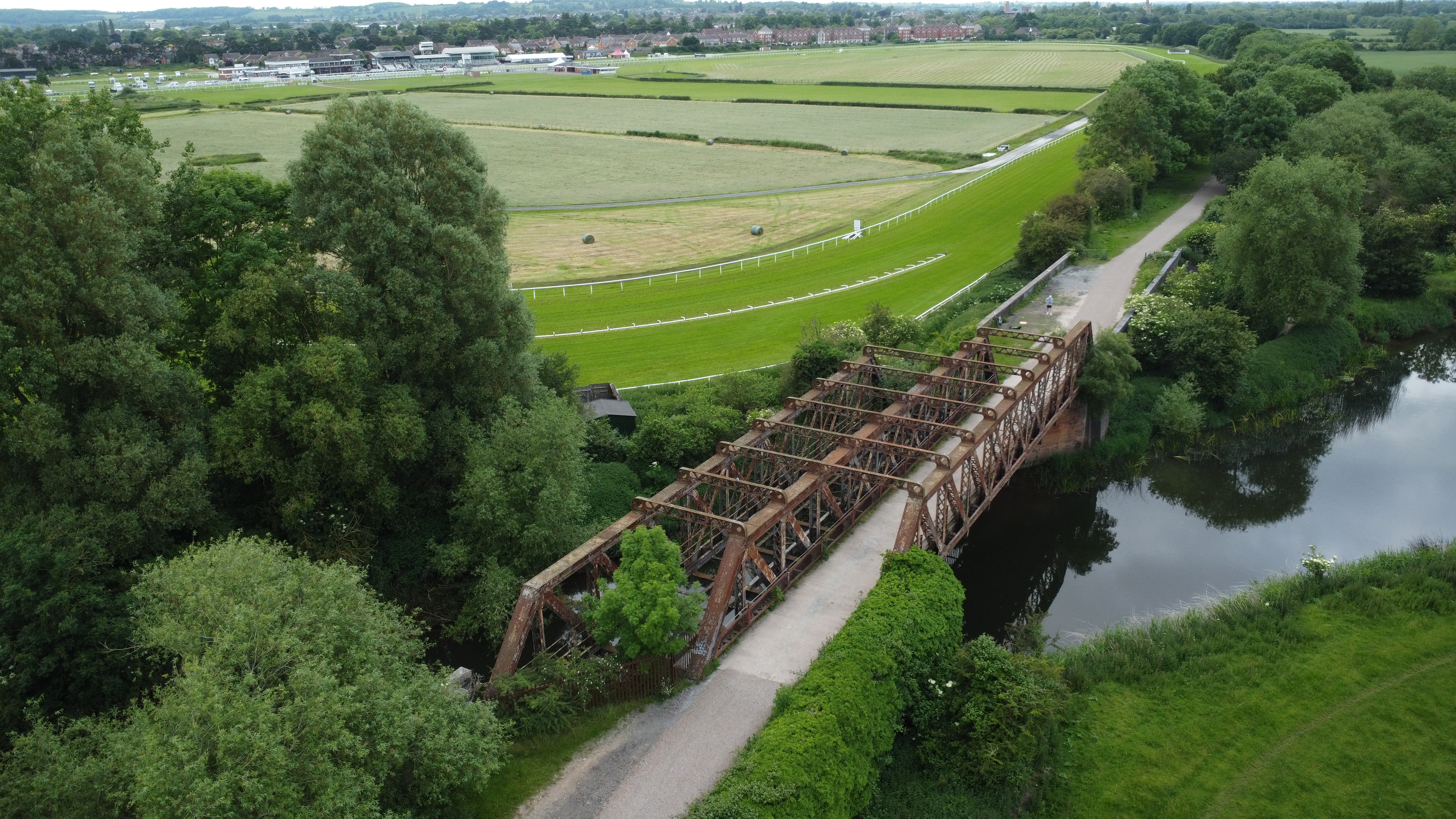 Aerial view of the Stratford-upon-Avon Greenway crossing a river on a historic iron bridge, with surrounding countryside, trees, and nearby racecourse visible.